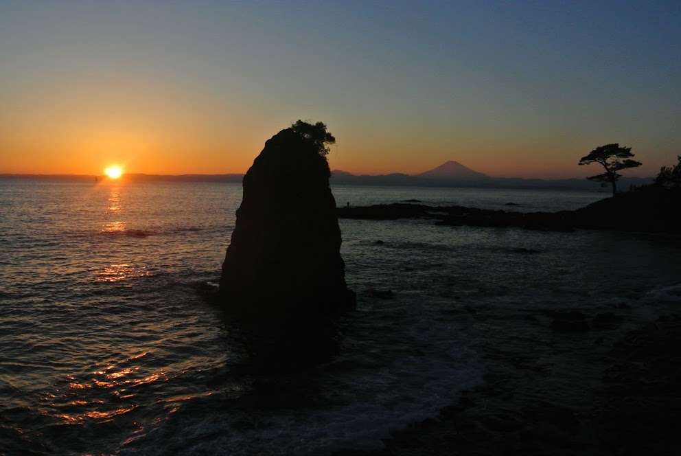 夕日の中、海に浮かぶ大きな石のシルエットが見える。