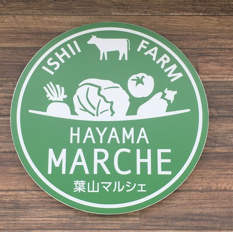 間山マルシェのロゴ。緑色の丸いロゴ。上半分には牛や野菜のデザインが、下半分には英語で葉山マルシェと書かれている。