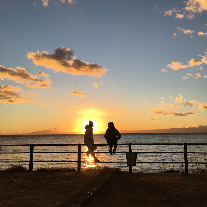 葉山公園・大浜海岸の夕日。公園の柵に腰掛ける若者2人の影が逆光となっている。