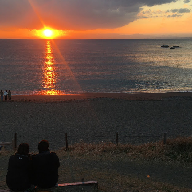 大浜海岸から見る夕日。奥紅色に光る夕日を、ベンチに座って眺めるカップルの後ろ姿が見える。